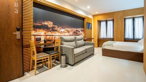 Quarto de hotel moderno com sofá, cama, mesa de jantar com cadeiras e uma janela grande com cortinas.