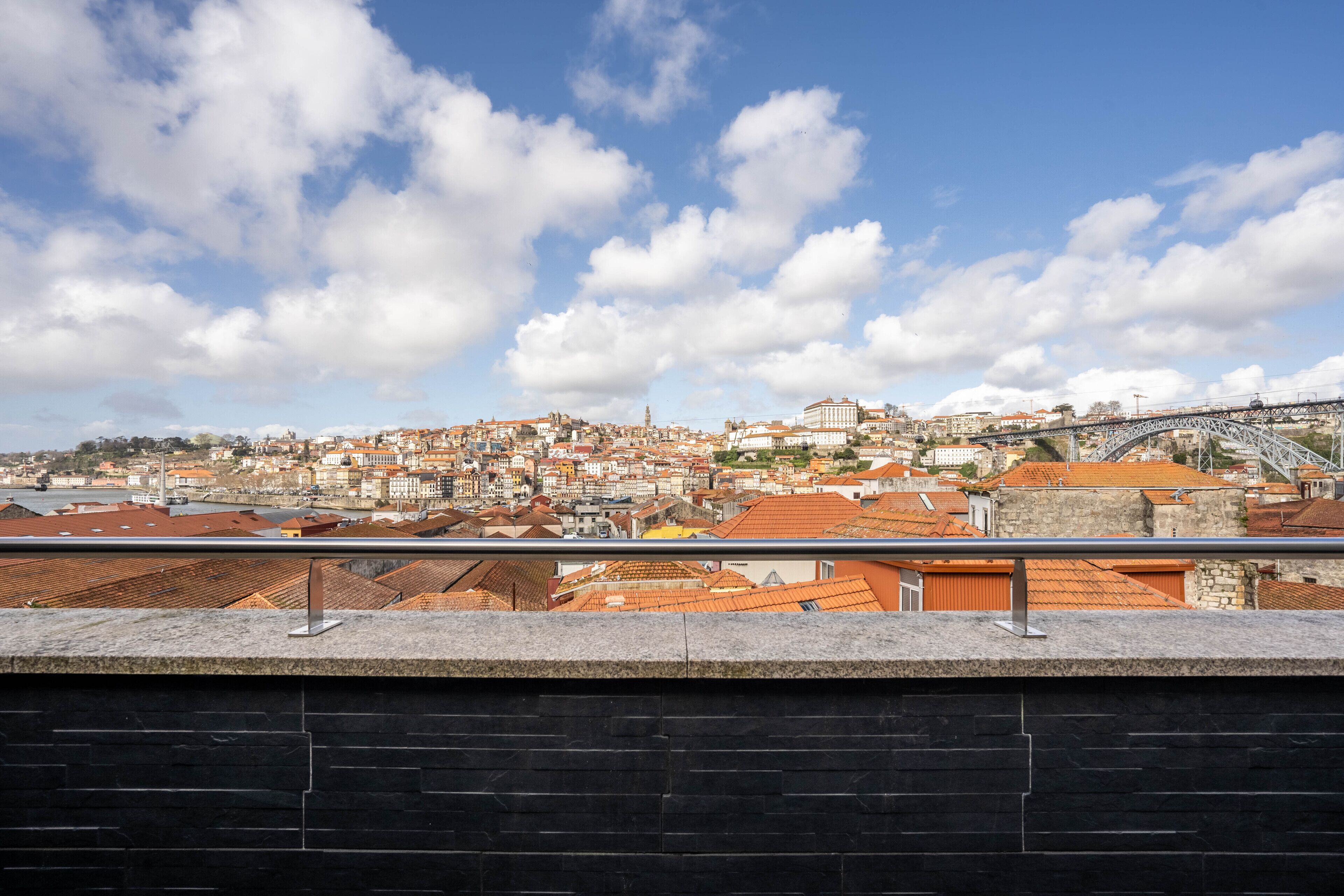 A panoramic view of a cityscape with a prominent bridge, terracotta-roofed buildings, and a clear sky.