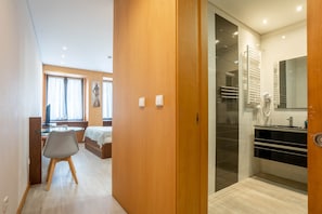 Habitación de hotel con puerta de madera, una cama, un escritorio y un baño con ducha y espejo.