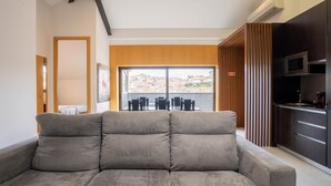 Uma sala de estar moderna com um sofá grande, uma área de cozinha com armários escuros e vista para um horizonte urbano através de amplas janelas.