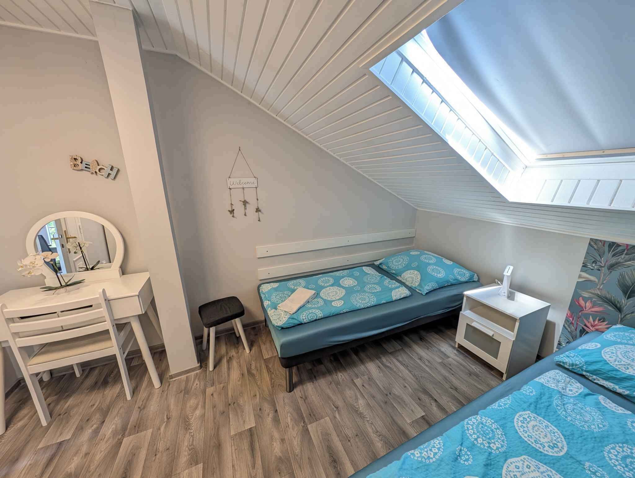 Un dormitorio con techo inclinado, una cama con ropa de cama estampada, una mesita de noche con lámpara, un escritorio pequeño con espejo y un adorno en la pared.