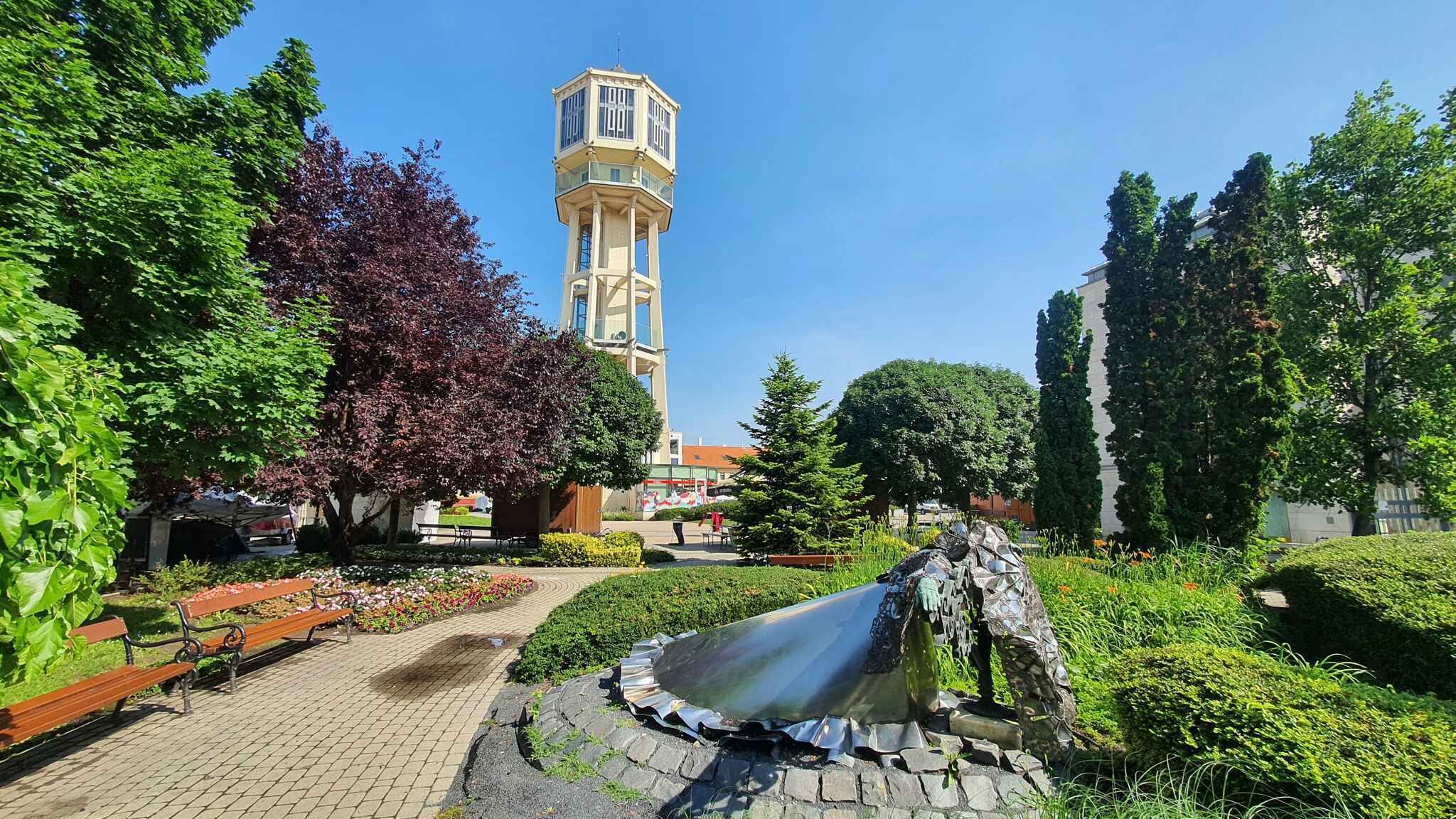 Un parque con fuente de agua, bancos y una torre al fondo.