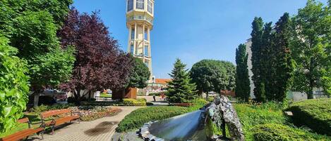 Un parque con fuente de agua, bancos y una torre al fondo.