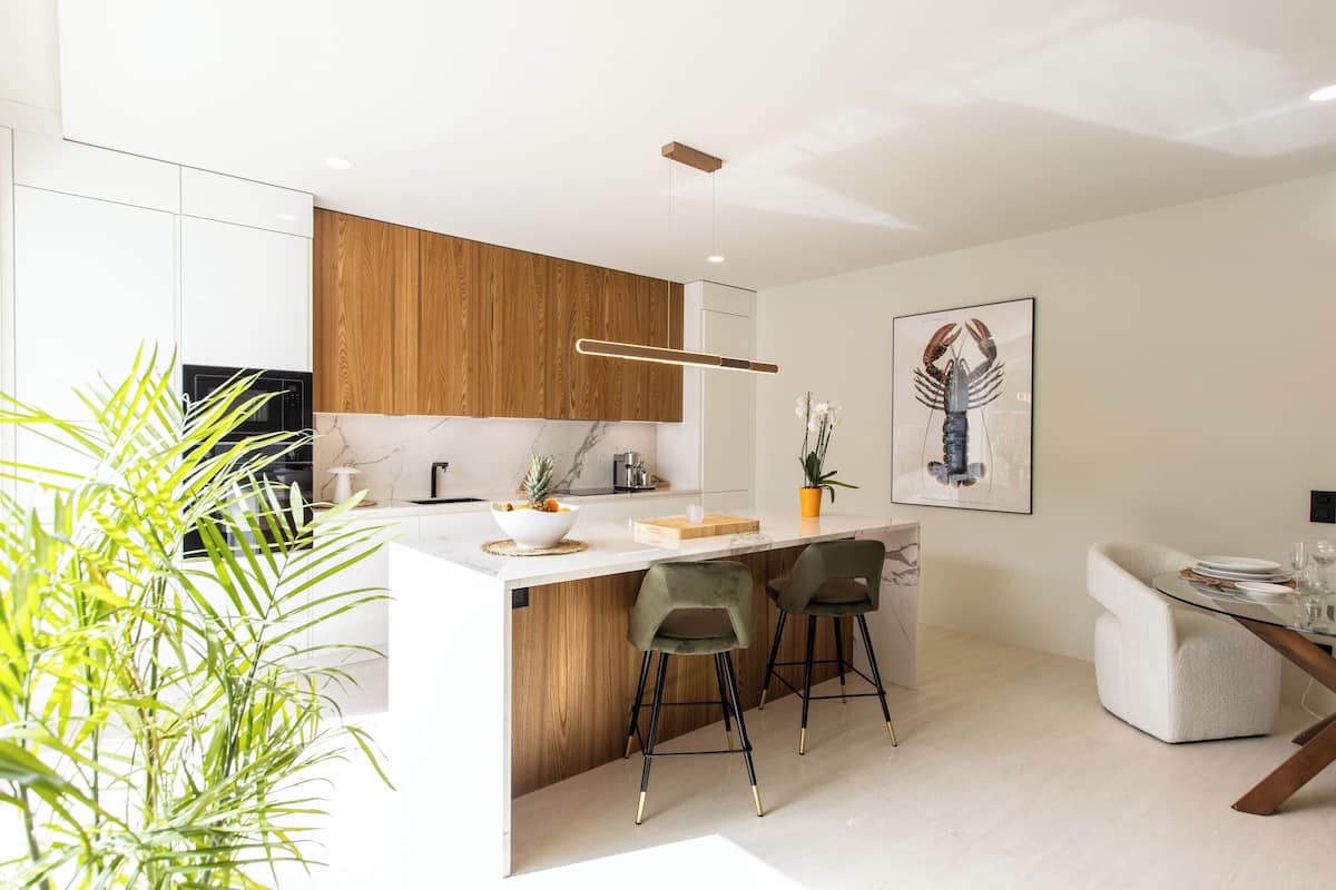 A modern kitchen with white cabinets, a wooden accent wall, a marble countertop, and a dining area with a sofa and bar stools.