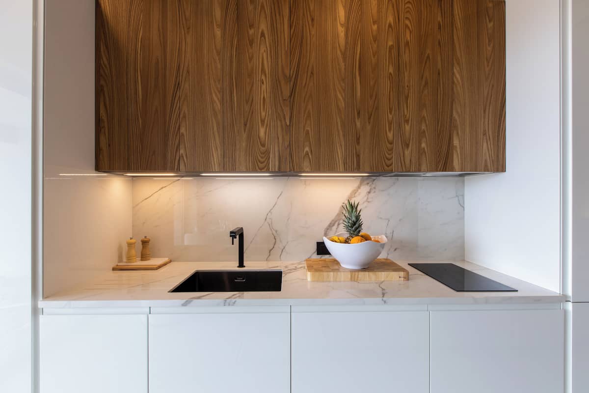 A modern kitchen with wooden cabinets, a marble countertop, a black sink, and a bowl of fruit.