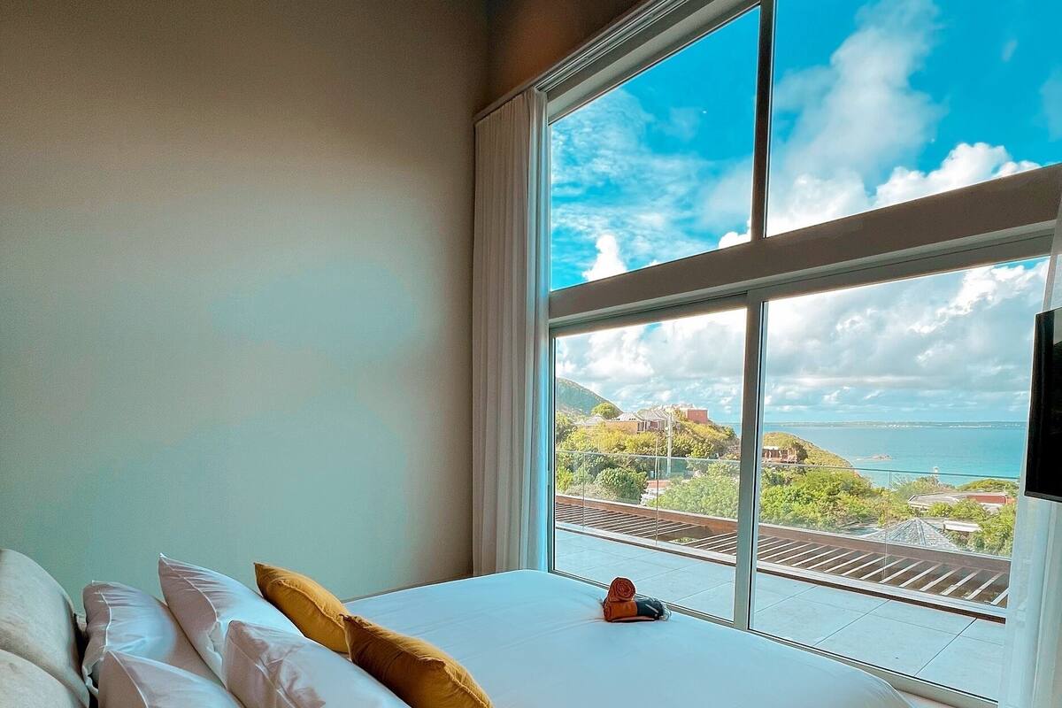 A bedroom with a large bed, white bedding, and a view of the sea through a large window.