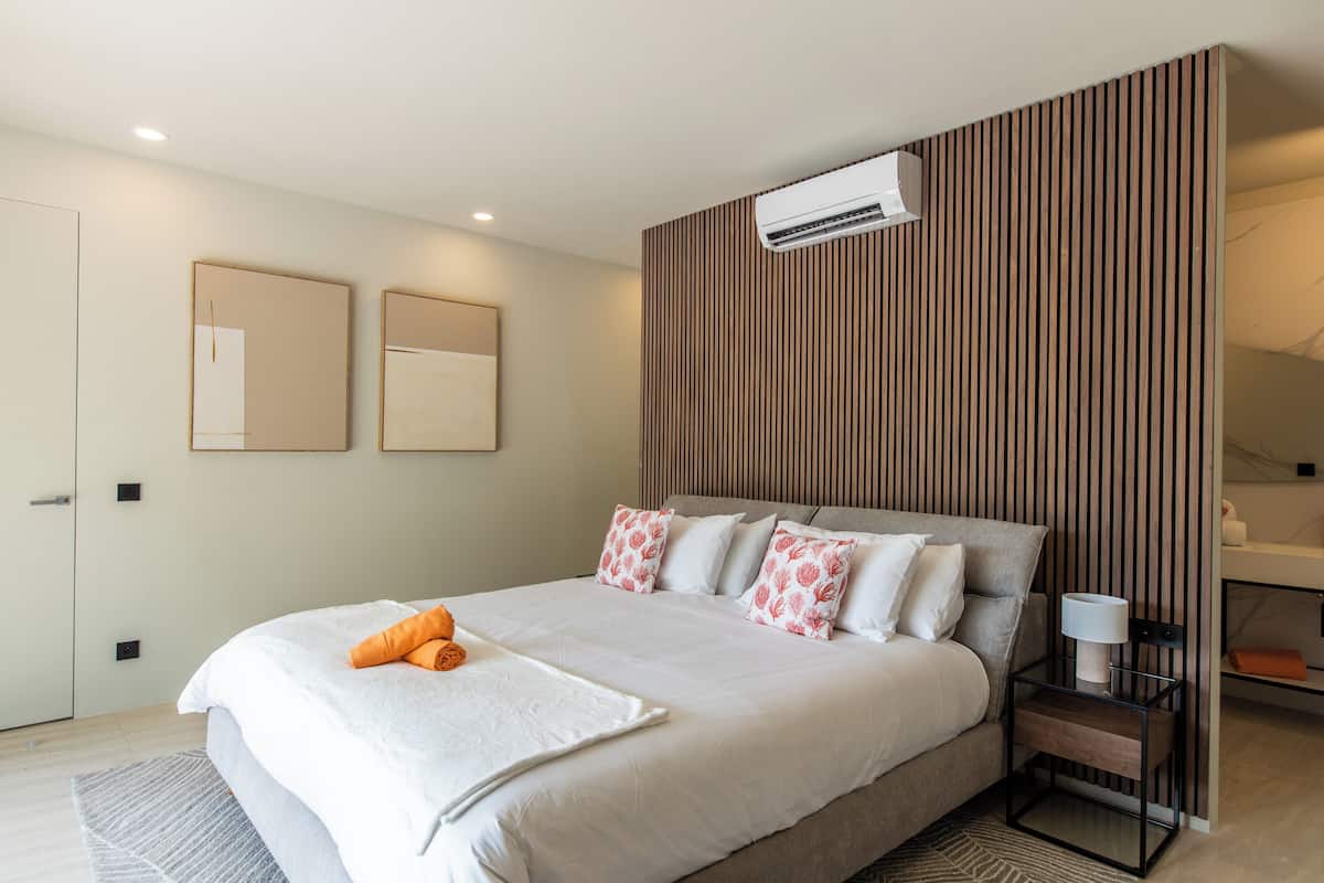 A modern bedroom with a large bed, a bedside table with a lamp, and a wall-mounted air conditioner.
