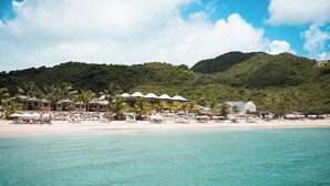 Un resort frente al mar con varias edificaciones tipo bungalow, una playa arenosa y colinas verdes exuberantes al fondo.