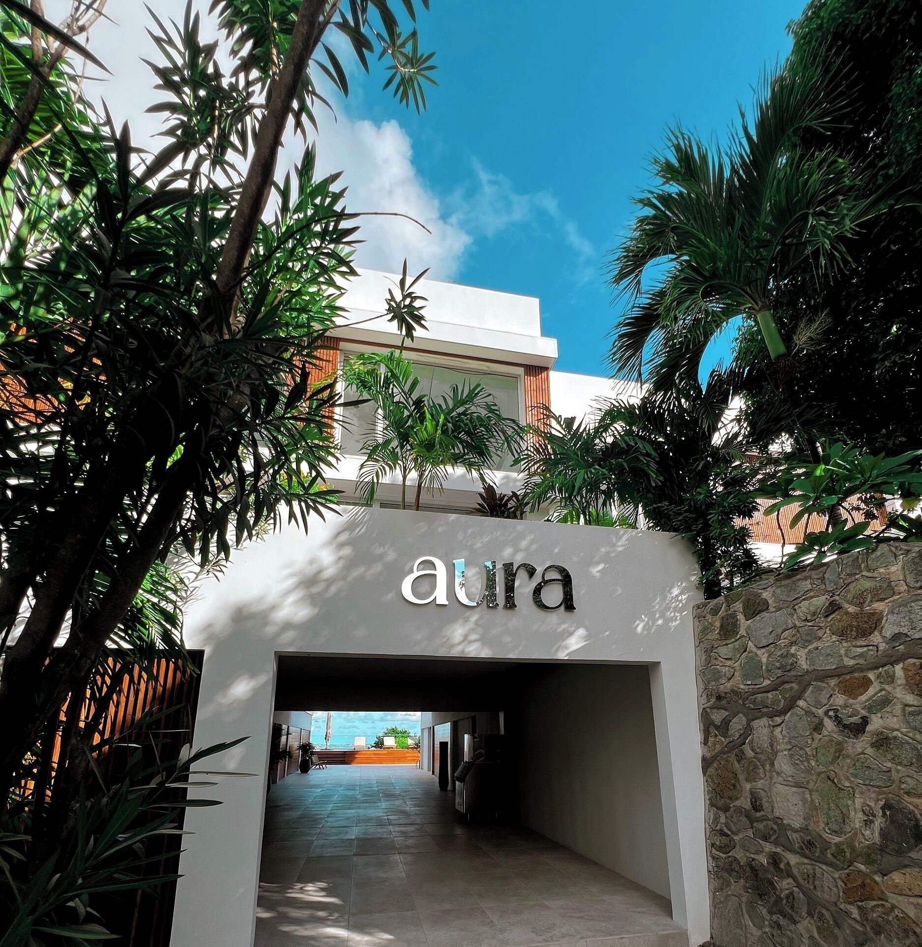 Entrada a un edificio llamado 'aura', con un muro de piedra y plantas tropicales.