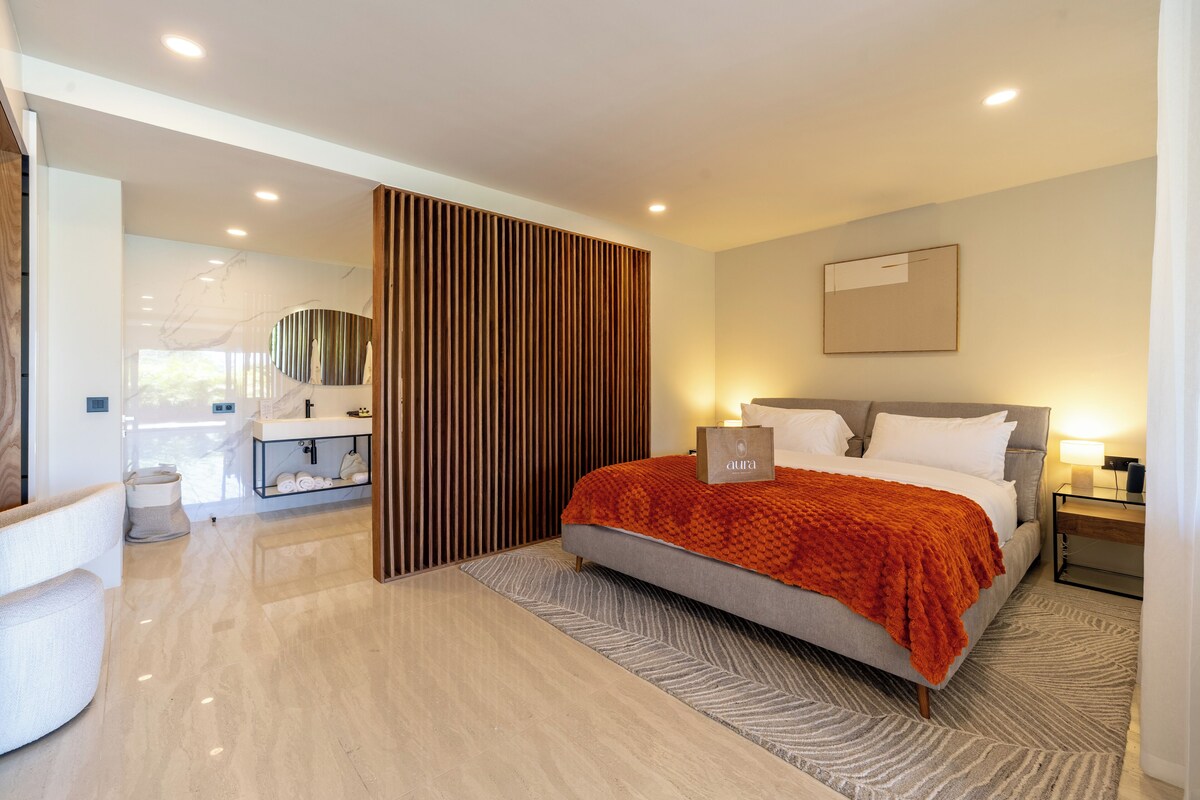 A modern hotel room with a large bed, a bedside table with a lamp, a bathroom with a mirror and sink, and a large window.