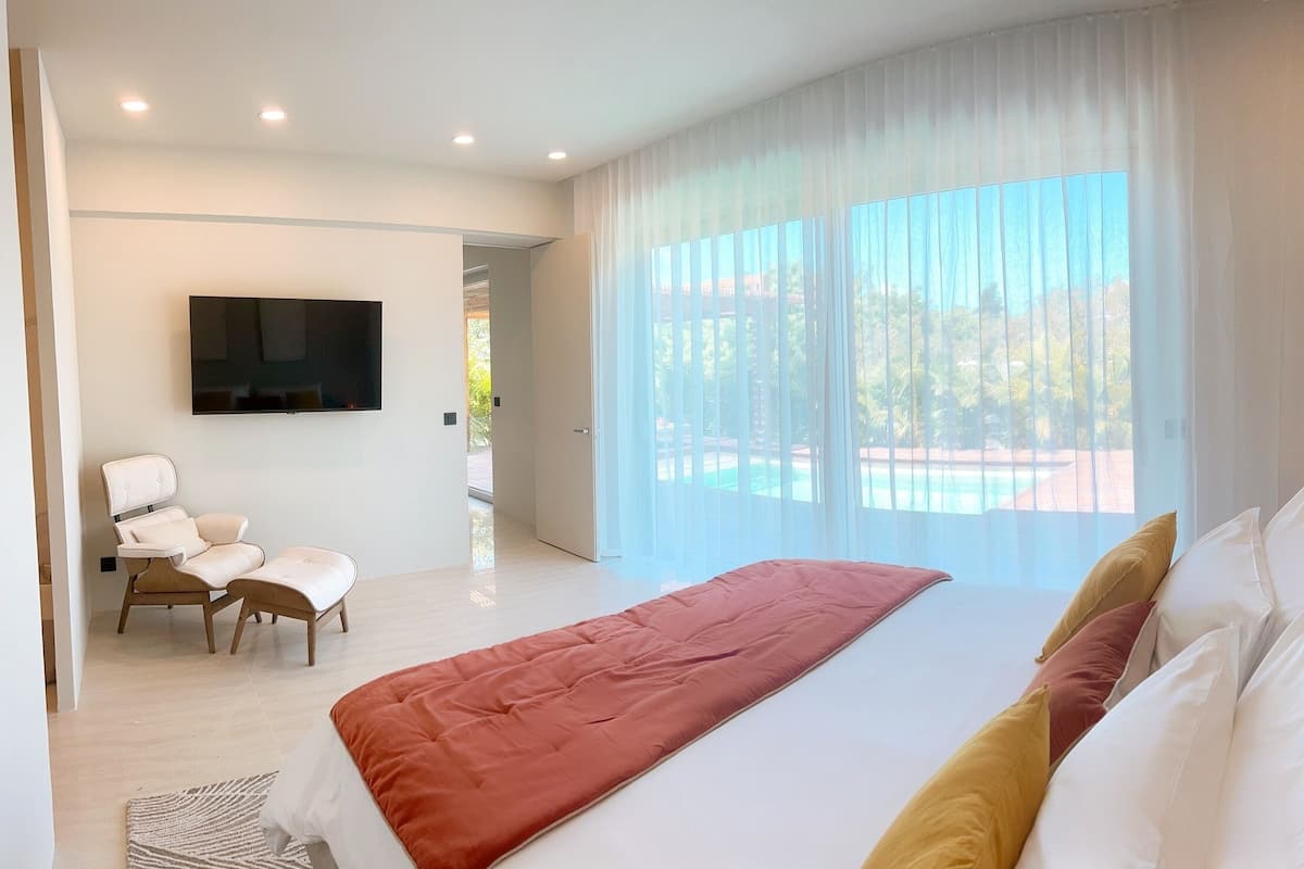 A modern hotel room with a large bed, a TV mounted on the wall, a seating area with a chair, and a view of the outdoors through large windows.