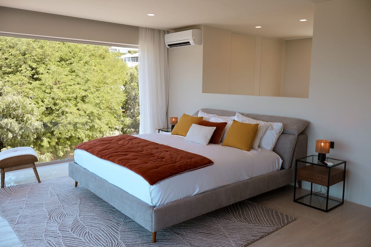 A modern bedroom with a large bed, a nightstand with a lamp, and a view of trees through a window.