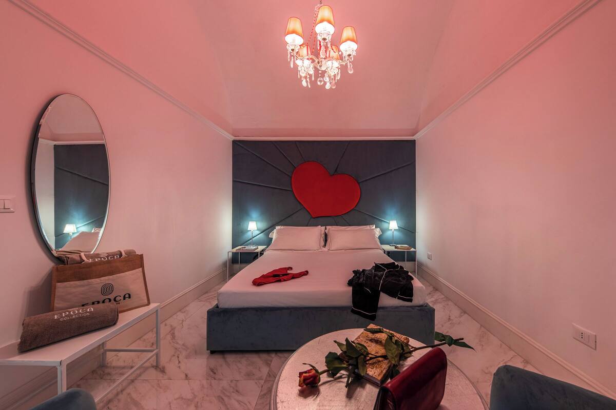 A bedroom with a bed, a large mirror, a chandelier, a heart-shaped decoration on the wall, and a bedside table with a lamp.