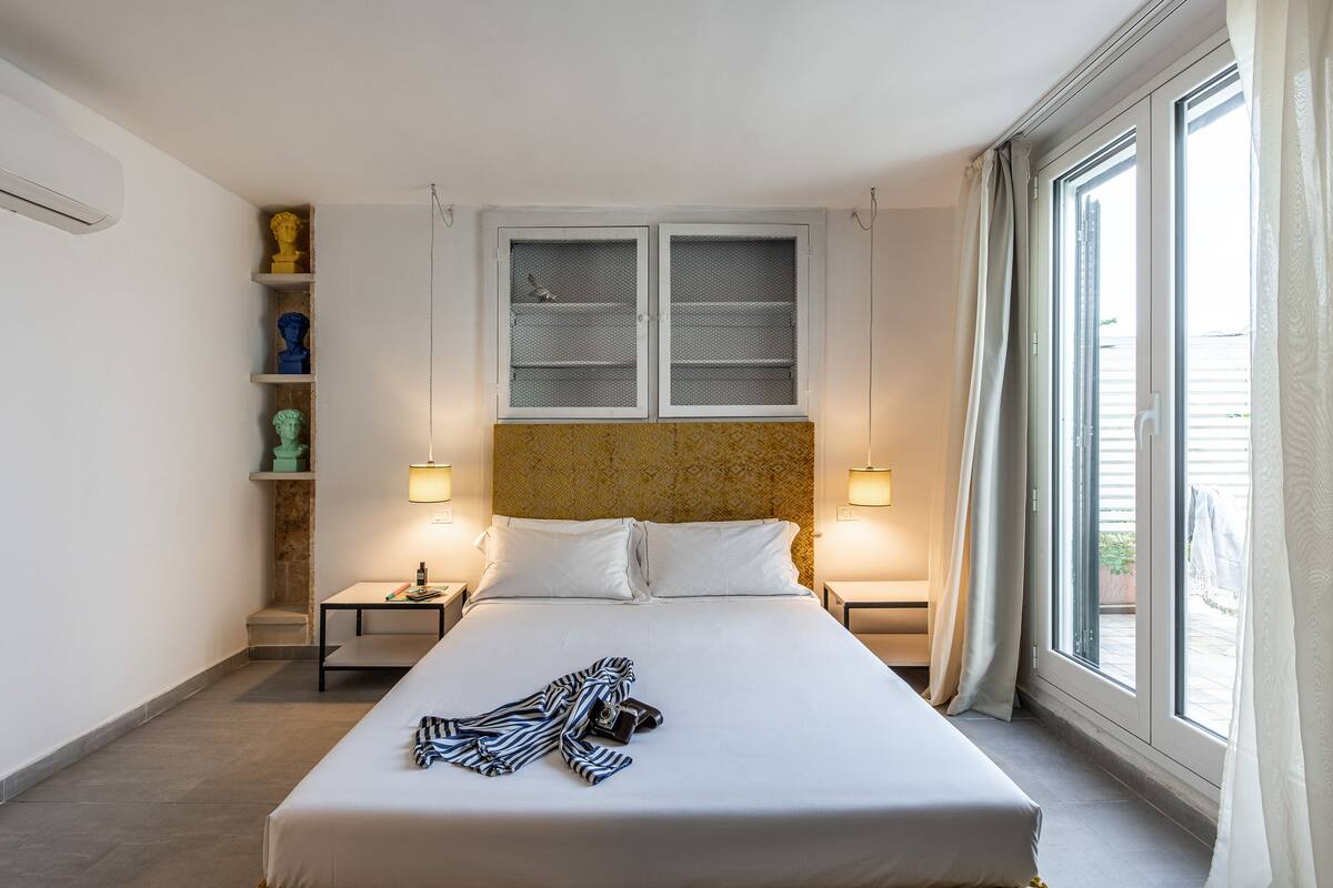A neatly made bed with a striped blanket and a camera, a small table with a lamp, and a shelf with decorative items.