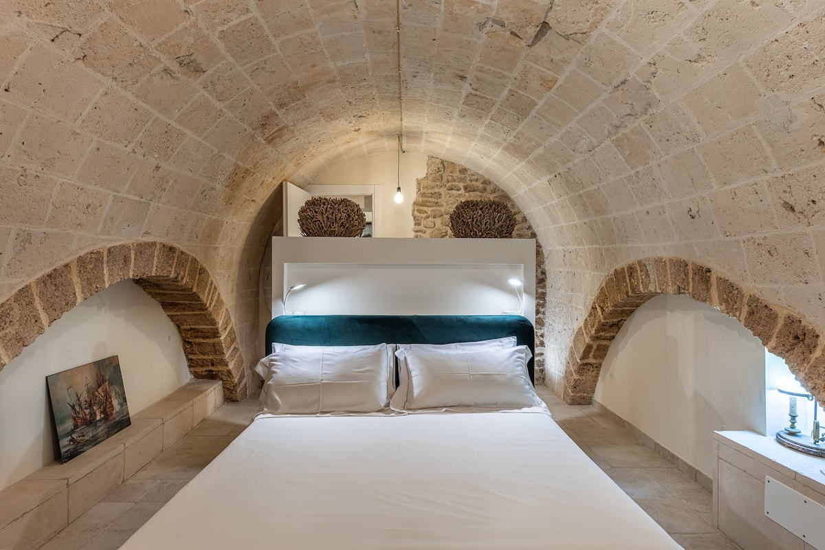 A bedroom with a stone arched ceiling, a bed with white linens, and a painting on the wall.