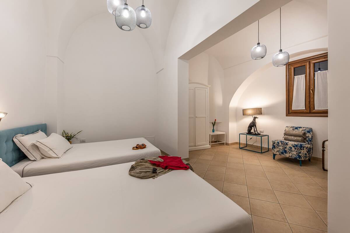 A hotel room with two beds, a chair, a small table, and a window with curtains.