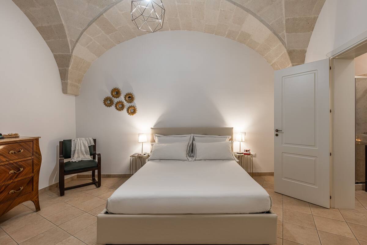 A bedroom with a large bed, a wooden dresser, a chair, and a door.