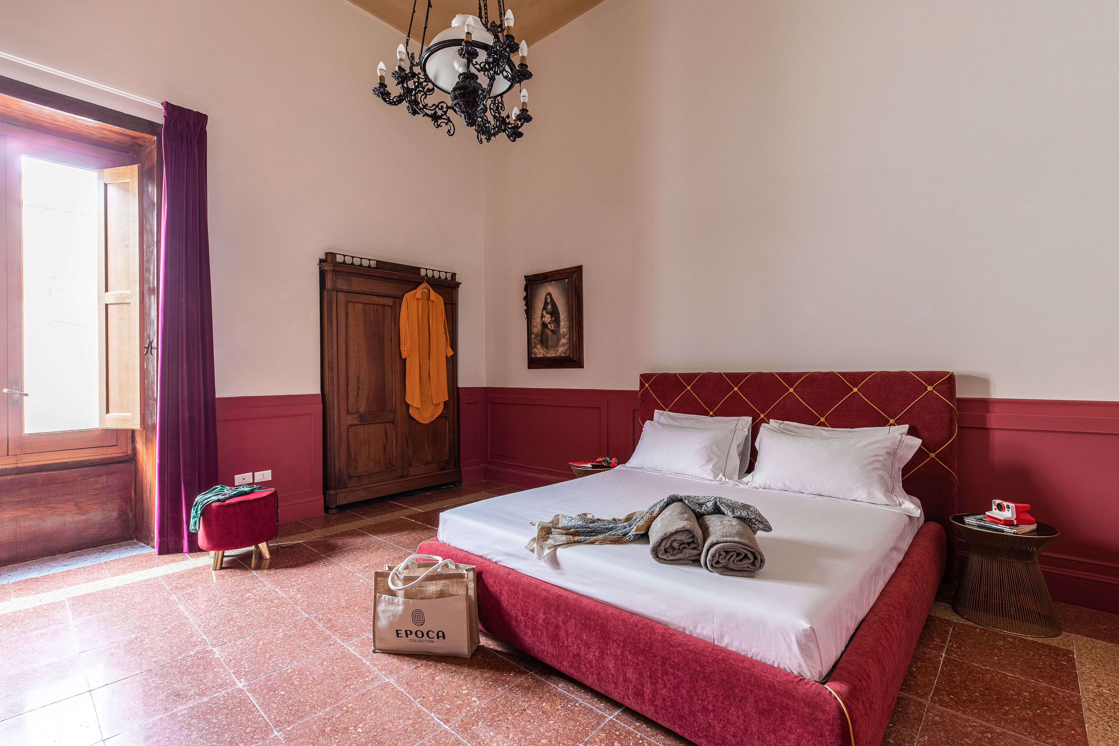 A hotel room with a bed, a wooden wardrobe, a chandelier, and a red accent wall.