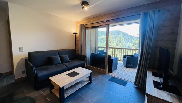 A modern living room with a sofa, coffee table, and a TV on a wooden stand. A balcony with a view of the outdoors is visible.