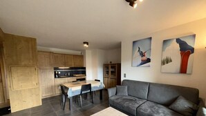 A modern living room with a grey sofa, a dining table with chairs, and a kitchen area with wooden cabinets.