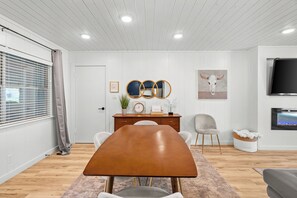 A modern living room with a wooden dining table, a grey sofa, a TV mounted on the wall, and a decorative bull skull painting.