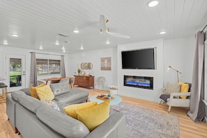 A modern living room with a grey sofa, yellow cushions, a flat-screen TV mounted on the wall, and a ceiling fan.