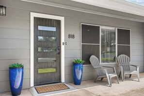 A house with a grey exterior, a front door with a number 818, two potted plants, and a set of chairs outside.
