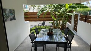 Uma área de refeições com mesa de vidro e cadeiras, cercada por um portão de madeira e uma bananeira grande.
