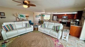 A living room with two white sofas, colorful pillows, a ceiling fan, and a kitchen area in the background.