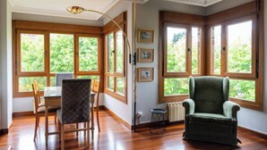A room with a dining table, chairs, a green armchair, and large windows with a view of greenery. - Apartamento Playa del Sardinero (Santander)