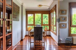 Un espace repas comprenant une table et des chaises, de grandes fenêtres donnant sur un paysage verdoyant et un luminaire au plafond.