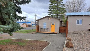 Un piccolo edificio a un piano con una porta gialla, un vialetto sterrato e un patio in legno con sedie.