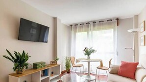 Ein Wohnzimmer mit Flachbildfernseher an der Wand, einem runden Glastisch, zwei Korbstühlen, einem Sofa mit rotem Kissen und einer kleinen Pflanze auf einem Holzregal.