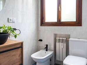 A bathroom with a white toilet, a bidet, a wooden vanity with a black sink, and a window above the bidet. - La casa del artista Sardinero (Santander)