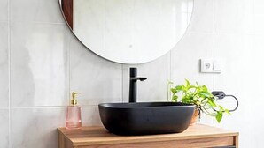 Un baño con un espejo redondo, un lavamanos tipo cuenco negro, un gabinete de madera, un dispensador de jabón rosa y una planta en maceta.