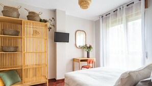 A bedroom with a bed, a wooden wardrobe, a desk with a chair, a television, and a window with sheer curtains.