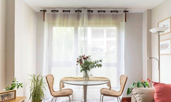 A room with a round table, two wicker chairs, a vase with flowers, a plant, and a window with sheer curtains. - La casa del artista Sardinero (Santander)