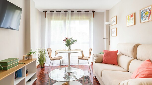 A living room with a sofa, a round glass coffee table, two wicker chairs, a dining table with a vase of flowers, and a flat-screen TV mounted on the wall.