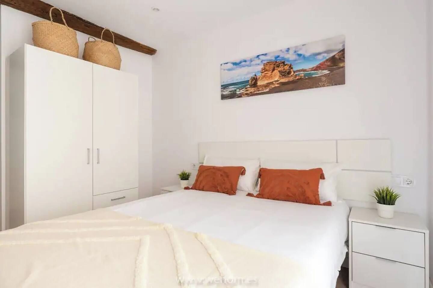 A bedroom with a bed, white wardrobe, and a painting of a coastal landscape.