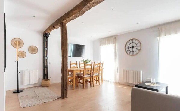 A spacious living room with a wooden ceiling beam, a dining area with wooden furniture, a clock on the wall, and a radiator. - Moderno y cómodo apartamento en el centro (Santander)