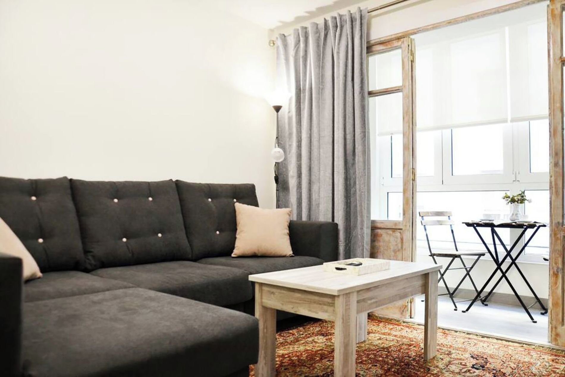 A modern living room with a dark grey sofa, a wooden coffee table, and a view of a balcony with a table and chairs.