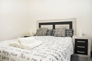 A neatly made bed with a white comforter and pillows with text, a bedside table with a lamp, and a nightstand with a small item on it. - Nuevo Apartamento con 3 dormitorios (Santander)