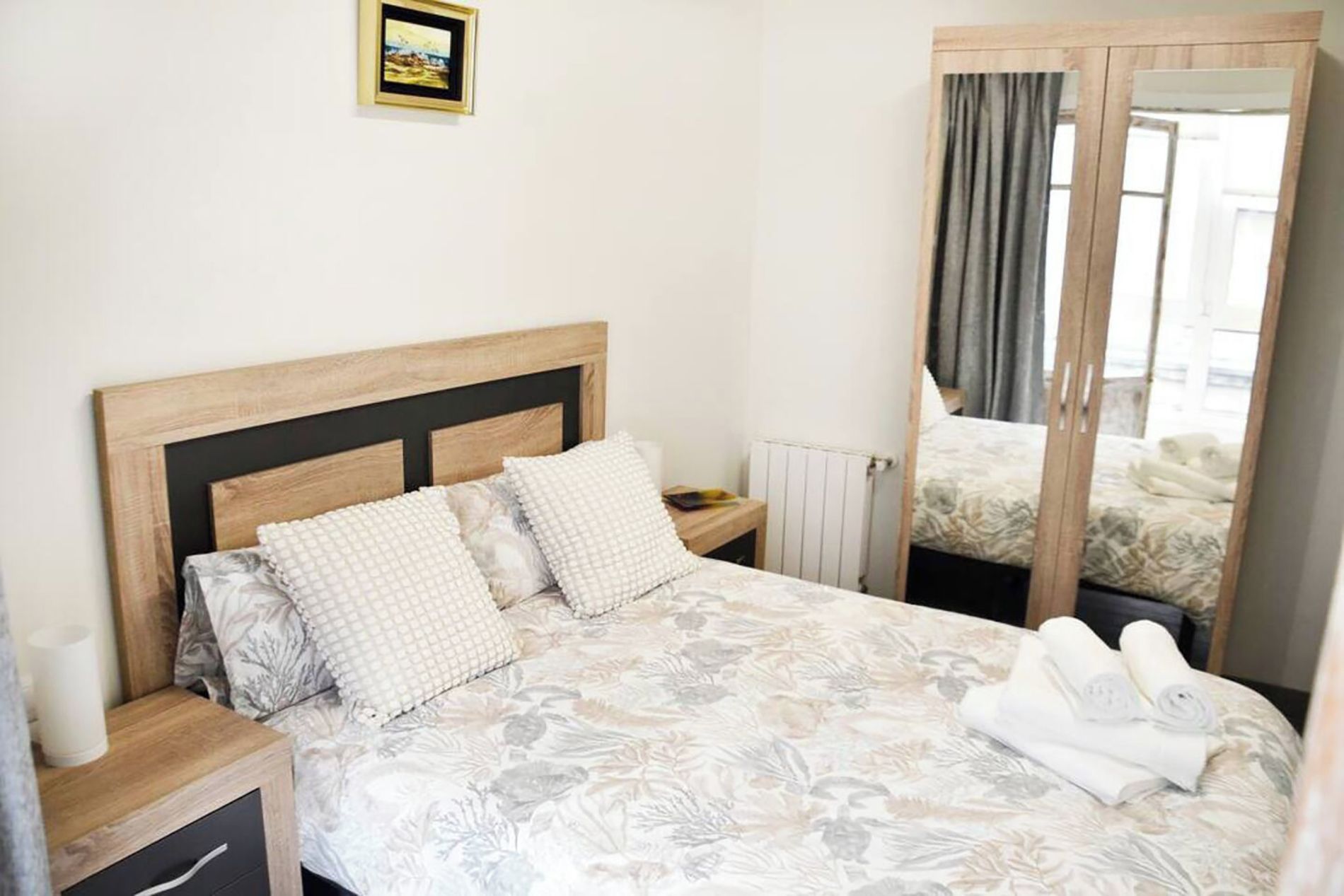 A neatly made bed with a floral bedspread, two pillows, and a wooden headboard. A small bedside table with a lamp and a book is visible. A mirror reflects part of the room.