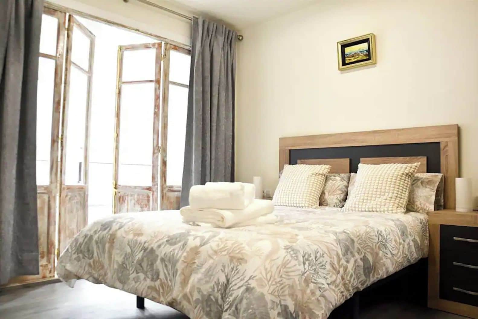 A bedroom with a bed, a nightstand, a framed picture on the wall, and a window with curtains.