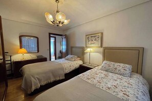 Habitación de hotel con dos camas, una araña, un escritorio con lámpara y una puerta que da a un balcón.