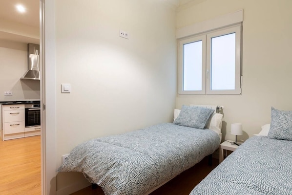 Una habitación pequeña y limpia con dos camas individuales, una ventana y una kitchenette al fondo.