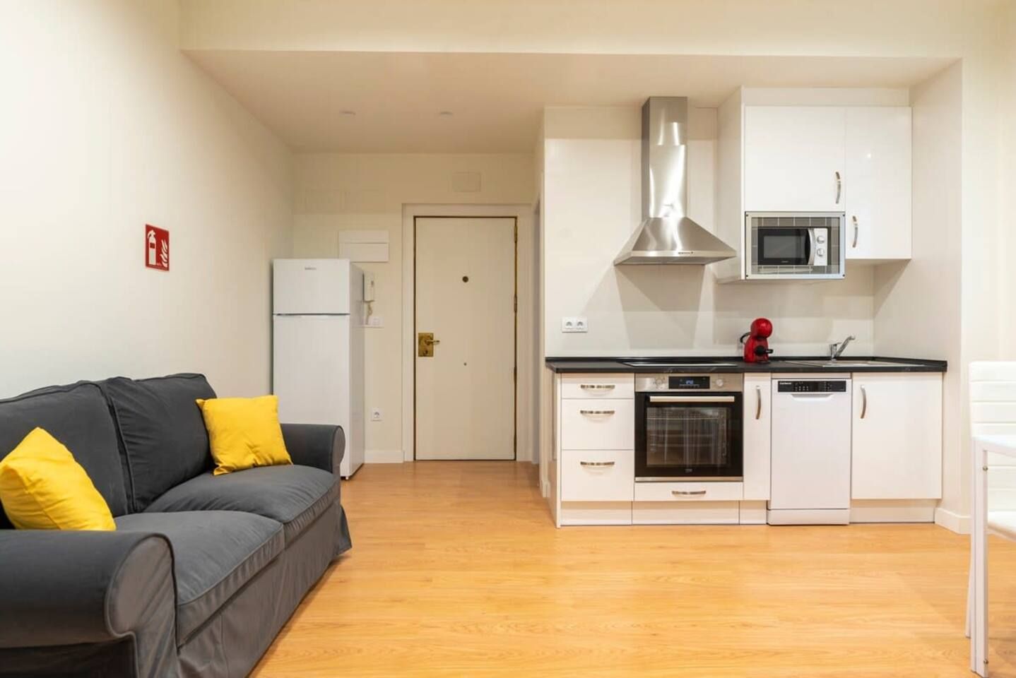 Un espacio habitable compacto con kitchenette, un sofá gris con cojines amarillos y suelo de madera.