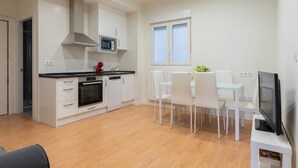 A modern kitchen with white cabinetry, a microwave, and a red kettle. There is a dining area with white chairs and a table, and a TV on a stand. - Casa junto a la Catedral (Santander)
