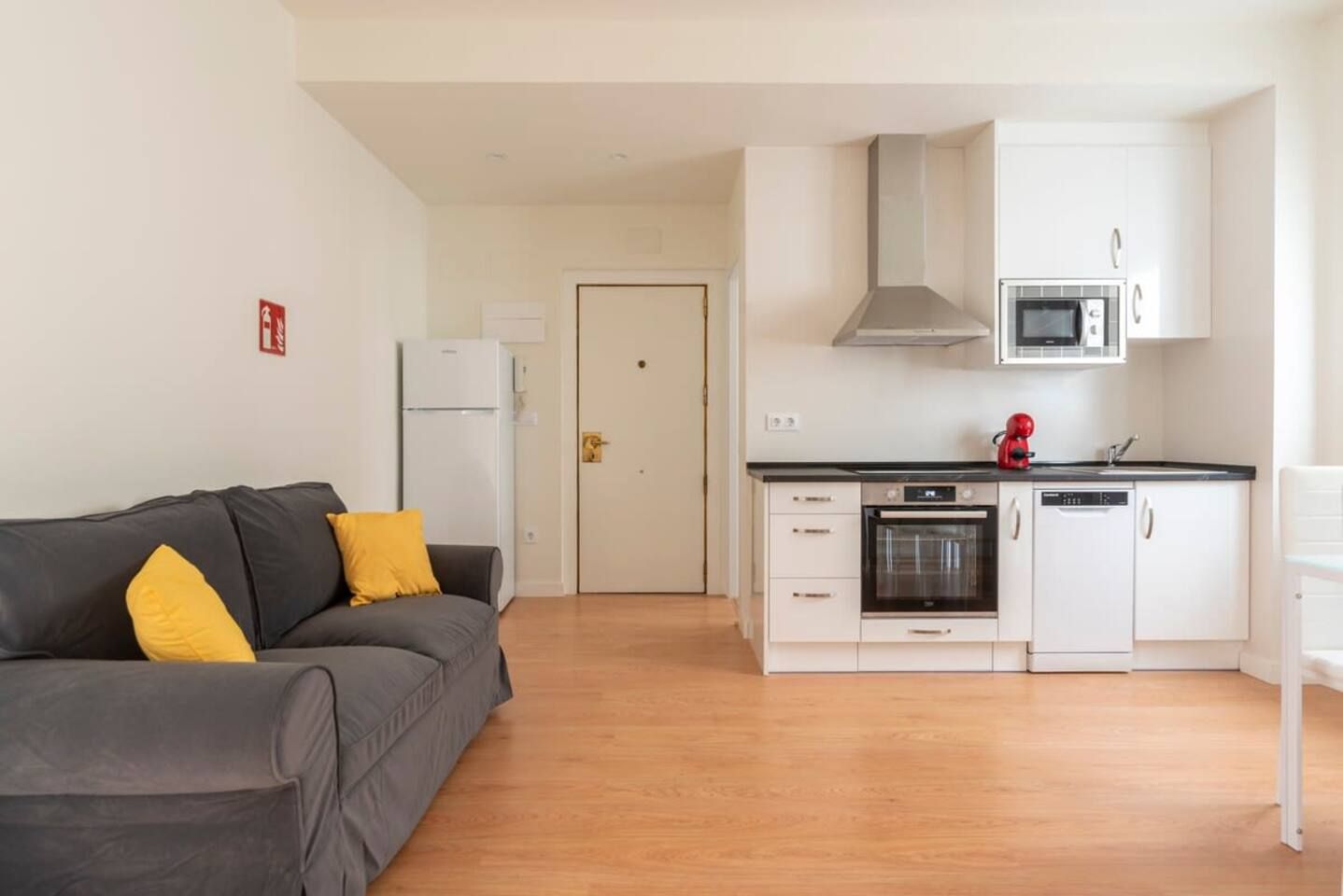 Un espacio habitable compacto con kitchenette, un sofá gris con cojines amarillos y una puerta blanca.