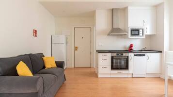 Un espacio habitable compacto con kitchenette, un sofá gris con cojines amarillos y una puerta blanca.